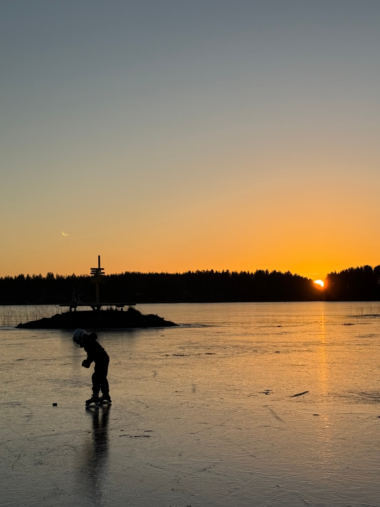 Luistelija jäällä ja aurinko laskemassa metsän taakse